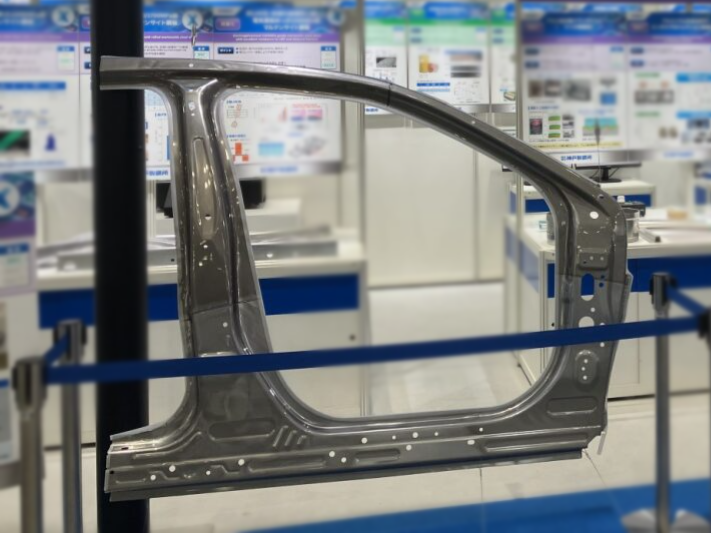 Metal car door frame on display in a factory or showroom, with a blue barrier in the foreground and a blurred background.