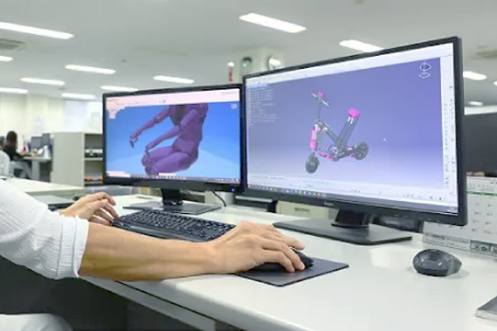 Person working at a desk with dual monitors displaying 3D CAD models.