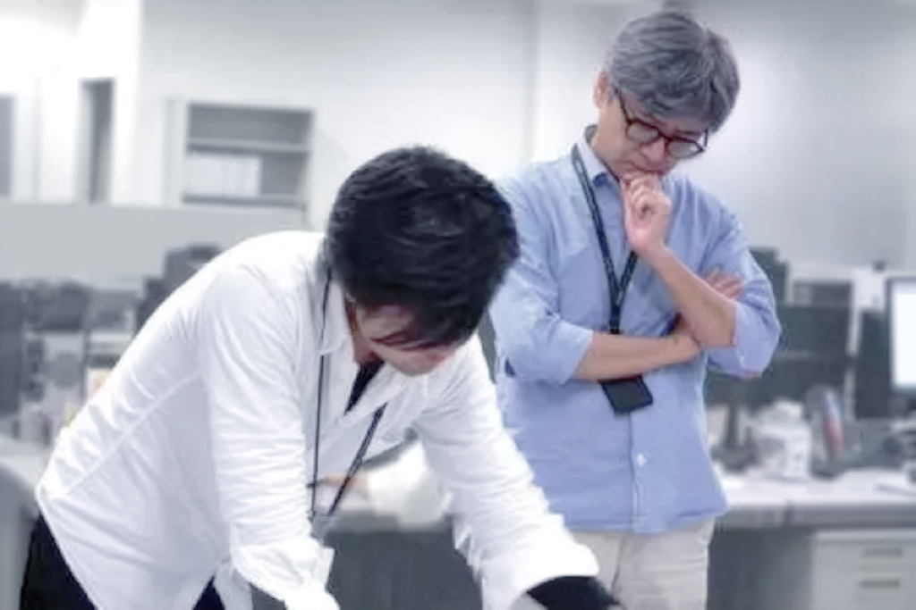 Two male coworkers in an office: one in a white shirt leaning over a desk, the other in a light blue shirt with glasses, arms crossed, pondering