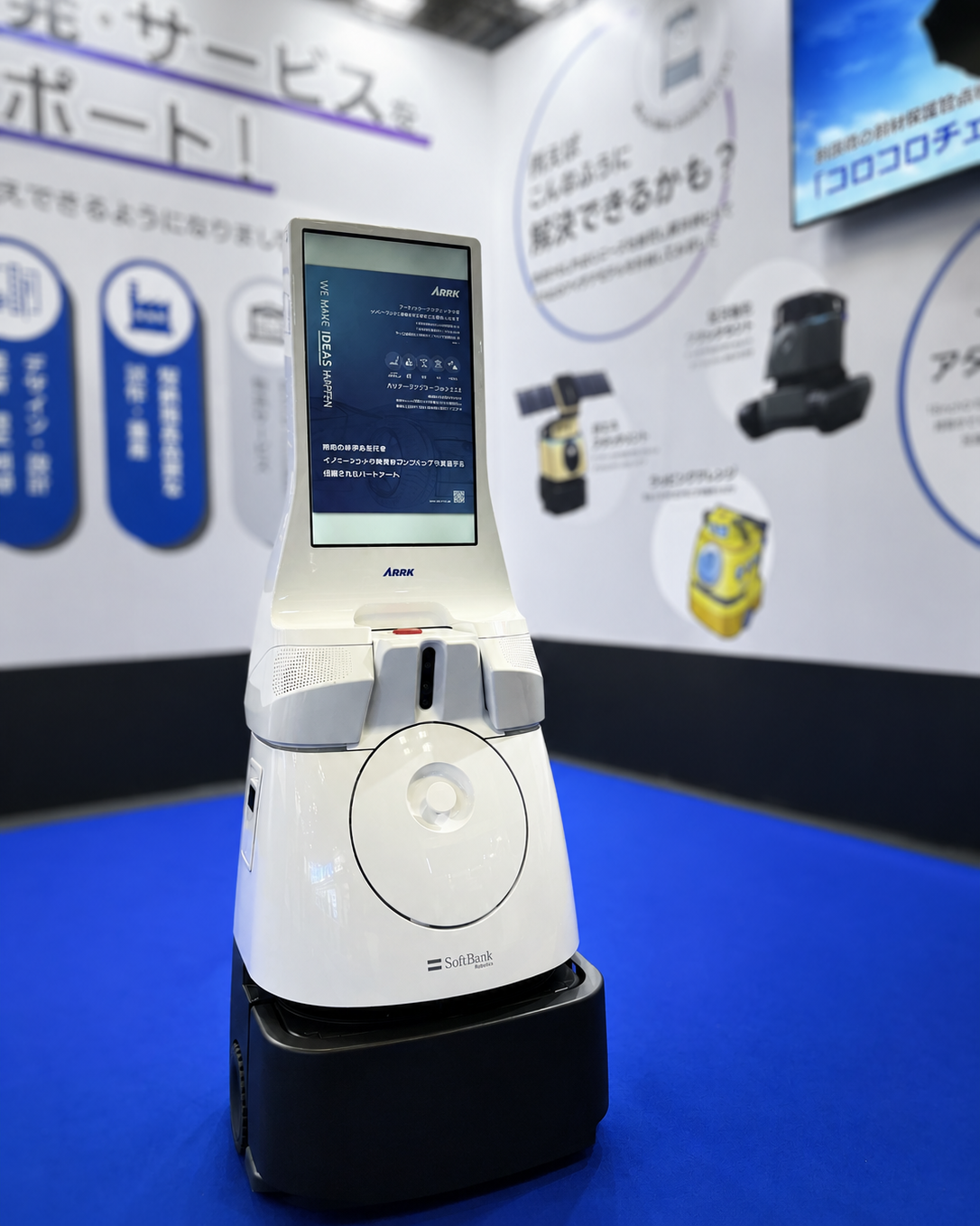 White service robot with a tall touchscreen display at a tech expo booth, standing on a black base on a blue floor.