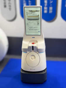 White self-service kiosk with a tall touchscreen displaying Japanese instructions and a red start button, mounted on a dark base on a blue surface.