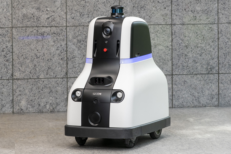 Autonomous white and black service robot on wheels with a forward-facing camera and blue LED ring, standing on a tiled outdoor surface.